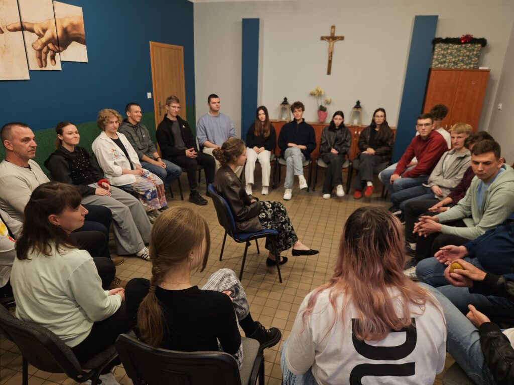 W jasnej sali z krzesłami ustawionymi w kręgu siedzi około dwudziestu osób w różnym wieku. Uczestnicy biorą udział w spotkaniu lub rozmowie grupowej.