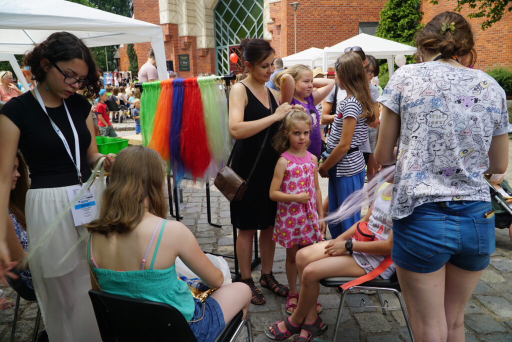 Dzieci i dorośli biorą udział w plenerowych warsztatach, gdzie kobiety zaplatają dzieciom kolorowe warkoczyki.”
