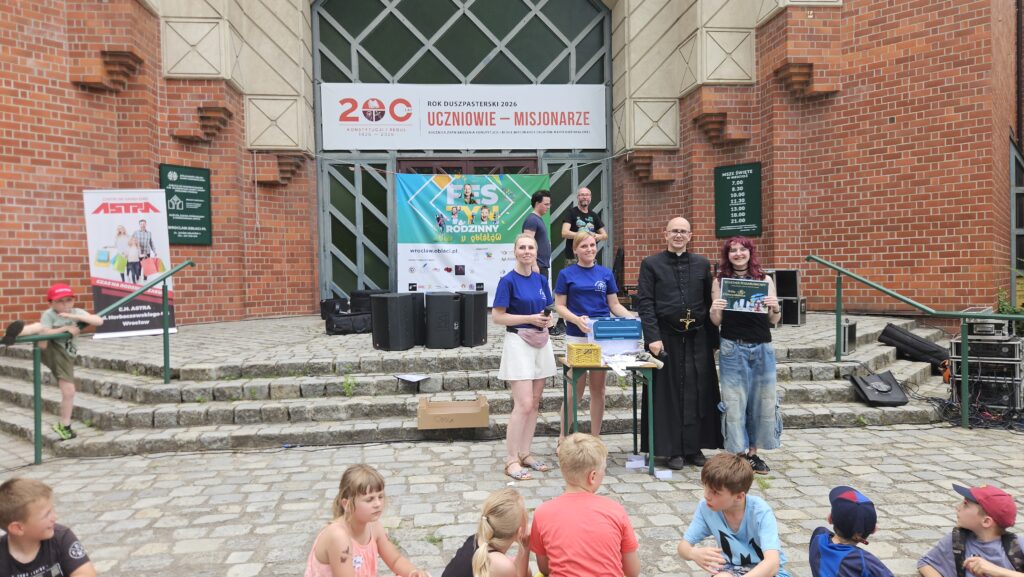 XI Festyn Rodzinny „U Oblatów” – oficjalne (i bardzo radosne!) podsumowanie Scena plenerowa przed wejściem do ceglanego kościoła. Na schodach stoją cztery osoby: dwie kobiety w niebieskich koszulkach, duchowny w czarnej sutannie oraz dziewczyna w szerokich jeansach trzymająca pamiątkowy dyplom. Nad nimi wisi baner z napisem „Uczniowie – Misjonarze”. Na brukowanym placu przed schodami siedzi grupa dzieci, tyłem do obiektywu, obserwując wydarzenie.