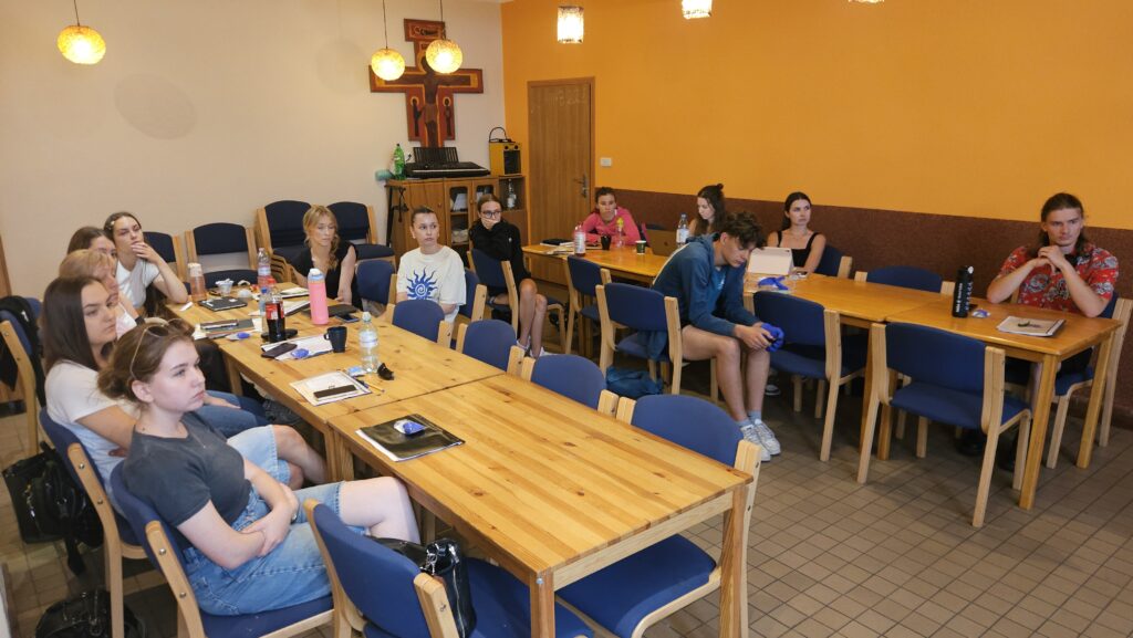 🎓 Weekendowy kurs zakończony! Kolejna ekipa gotowa na wakacyjne wyzwania Grupa uczestników kursu siedzi przy drewnianych stołach ustawionych w podkowę w sali szkoleniowej. Na ścianie wisi duży drewniany krzyż, a nad stołami wiszą lampy. Uczestnicy robią notatki i słuchają prowadzącego, tworząc atmosferę skupionej pracy.