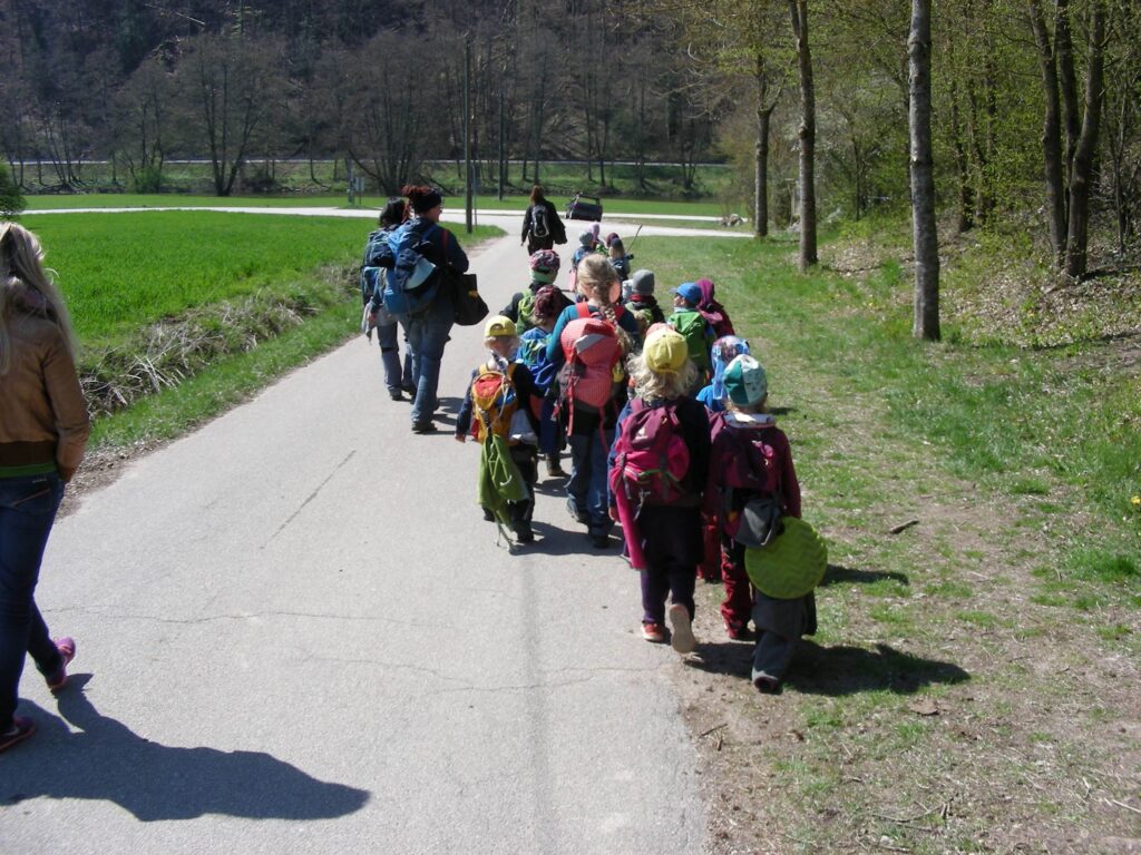 Grupa dzieci idzie asfaltową ścieżką w terenie zielonym, obok nich kilku dorosłych opiekunów. Dzieci mają kolorowe ubrania i plecaki. Po lewej stronie widać trawę, po prawej drzewa, a w tle zalesione wzgórze. Scena przedstawia spacer lub wycieczkę w plenerze.
